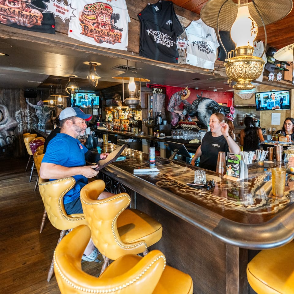 A man sits at the Krazy Kooters bar, while a bartender smiles and other patrons are visible.
