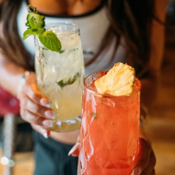 Two hands holding up two tall, refreshing cocktails, one yellow with mint, one red with pineapple.