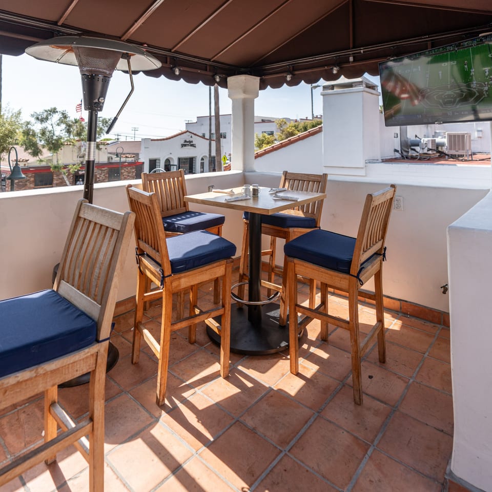 Krazy Kooters' outdoor patio with high wooden tables and chairs, blue cushions, and a TV showing sports.