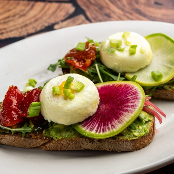 Two avocado toasts with poached eggs, watermelon radish, sun-dried tomatoes, and green onions.