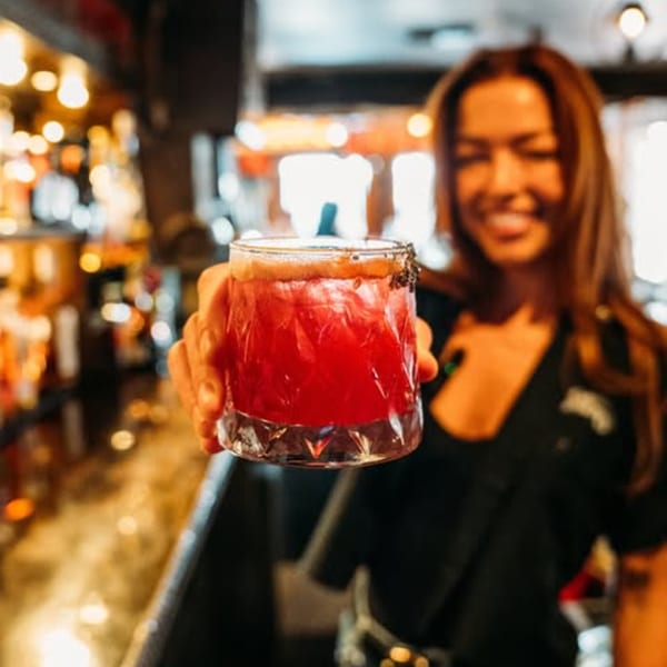 A smiling bartender at Krazy Kooters holds out a vibrant red cocktail with a salted rim.