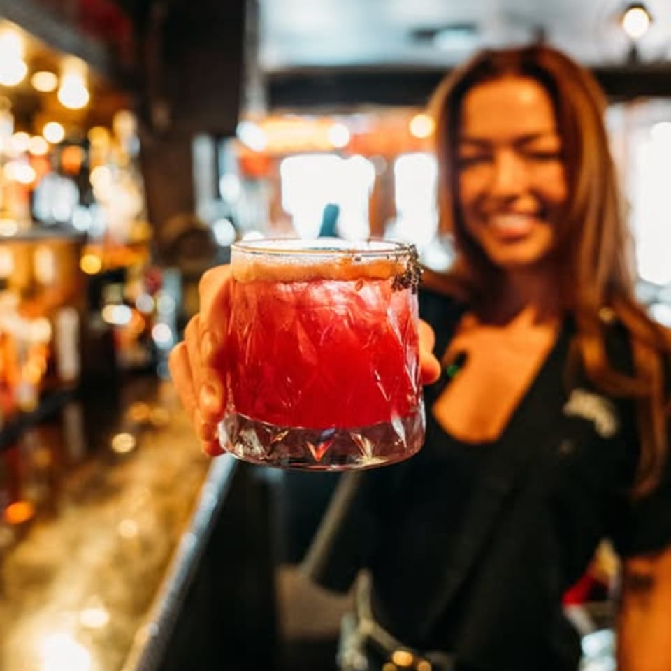 A smiling bartender at Krazy Kooters holds out a vibrant red cocktail with a salted rim.