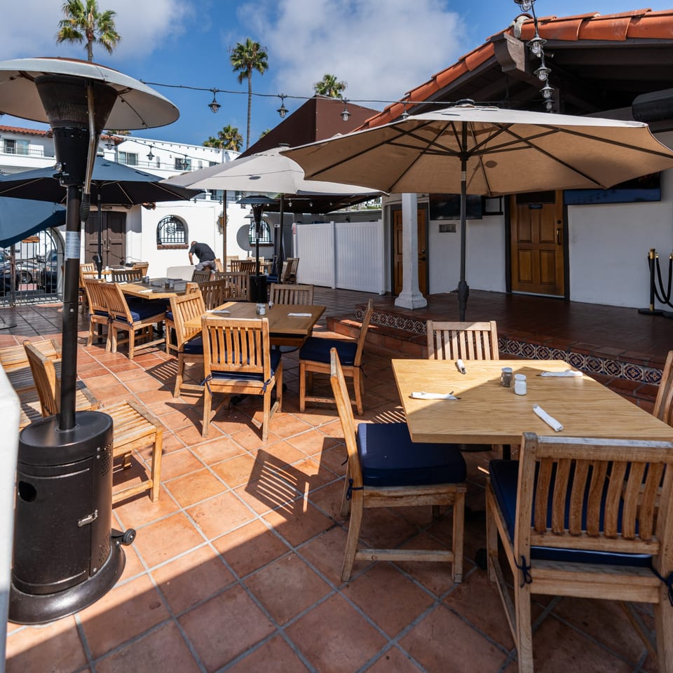 Krazy Kooters' outdoor patio with wooden tables, blue cushioned chairs, umbrellas, and heaters under palm trees.